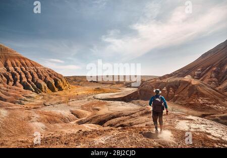 Tourist with backpack walking at the dusty trail on surreal red mountains against blue sky in desert park Altyn Emel in Kazakhstan Stock Photo