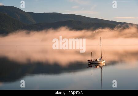 A lone sailboat is anchored on the glass-like calm water in a cove surrounded by mountains at sunrise. Stock Photo