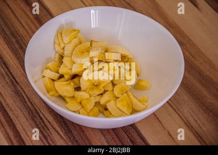 White bowl with small yellow banana pieces on a wooden table Stock Photo