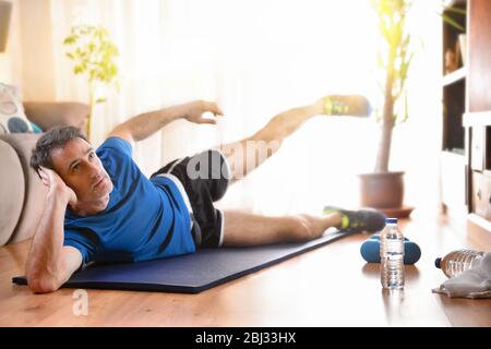 Man doing leg exercises lying on mat in his living room with water and dumbbells on the floor