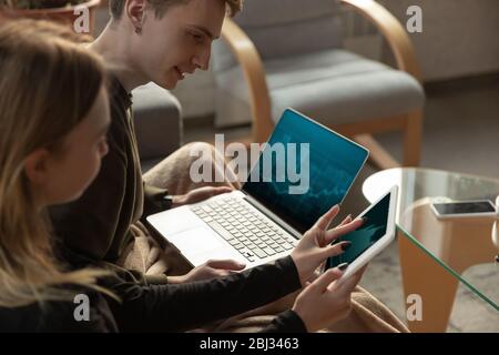 Attractive young couple using devices together, tablet, laptop, smartphone, headphones wireless. Communication, gadgets concept. Technologies connecting people in self-insulation. Lifestyle at home. Stock Photo