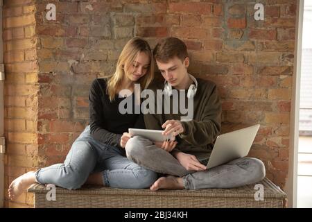 Attractive young couple using devices together, tablet, laptop, smartphone, headphones wireless. Communication, gadgets concept. Technologies connecting people in self-insulation. Lifestyle at home. Stock Photo