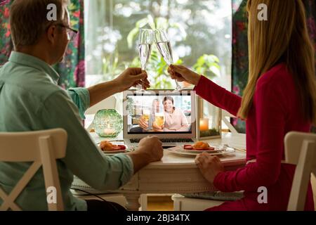 Man and woman sitting at the dining table, having dinner, drinking Champagne and having video call with senior parents on laptop. Staying home Stock Photo