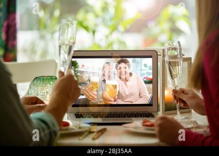 Man and woman sitting at the dining table, having dinner, drinking Champagne and having video call with senior parents on laptop. Staying home Stock Photo