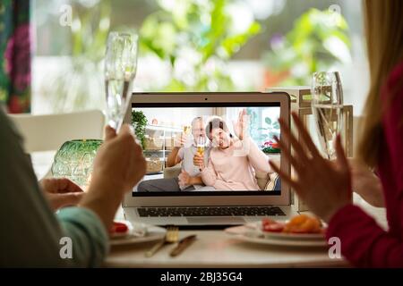 Man and woman sitting at the dining table, having dinner, drinking Champagne and having video call with senior parents on laptop. Staying home Stock Photo
