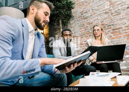 Making great decisions. Young Caucasian blond woman and two men, Caucasian and African, gesturing and discussing something with smile sitting at the Stock Photo