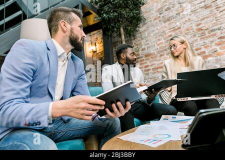Making great decisions. Young Caucasian blond woman and two men, Caucasian and African, gesturing and discussing something with smile sitting at the Stock Photo