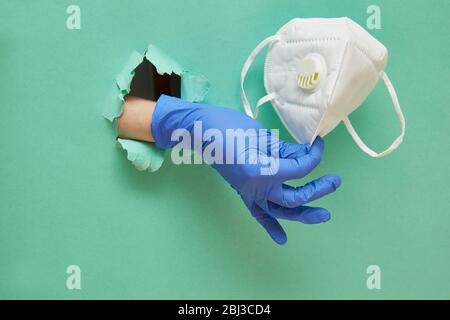 A hand in a protective blue glove holds a mask to protect the respiratory system and face. Hole in paper with copy space. Green background. Stock Photo