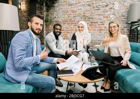 Successful excited multi-ethnic group of business people sitting together in office around the table with papers and laptop, showing thumbs up gesture Stock Photo