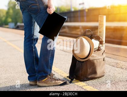A man waiting for the train with green travel  rucksack, hat, maps, glasses and holding the tablet in his hand at the empty train station during quara Stock Photo