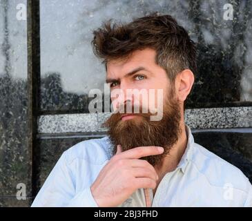 Man with beard and mustache on thoughtful face, black marble background. Masculinity concept. Hipster with tousled hair touches beard while looking into distance. Guy looks interested and curious Stock Photo