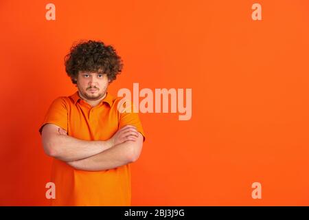 Serious with hands crossed. Caucasian young man's monochrome portrait on orange studio background. Beautiful male curly model in casual style. Concept of human emotions, facial expression, sales, ad. Stock Photo