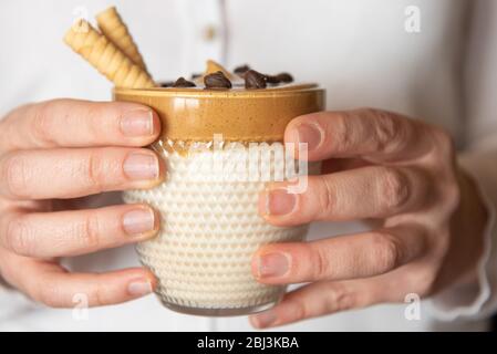 Female hands hold homemade dalgon coffee in a glass with milk on a wooden stand. Trendy fluffy creamy whipped coffee. Stock Photo