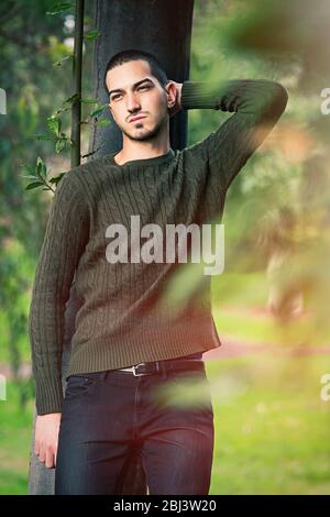 Young and handsome guy leaning against a tree in nature relaxing in a natural park. Autumn fashion. Stock Photo