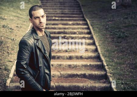 Model handsome man near steps. A handsome young man model outdoors. Near the steps in a park. He wears a leather jacket. Short hair almost shaved. Exp Stock Photo