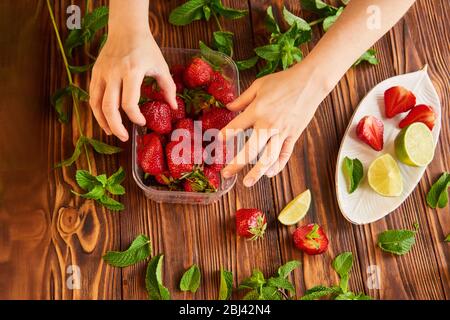 woman picks strawberries. berries in a plastic container. lime, strawberry, mint - ingredients for making lemonade on brown wooden table Stock Photo