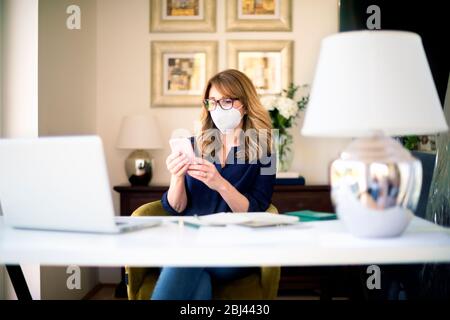 Portrait shot of middle aged woman wearing face mask while working at home on her notebook and text messaging during coronavirus pandemic. Stock Photo
