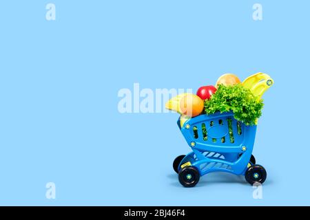 A grocery shopping cart full of salad, oranges, bananas, apples and bread. Close-up concept with place for text on a blue background. For articles, ad Stock Photo