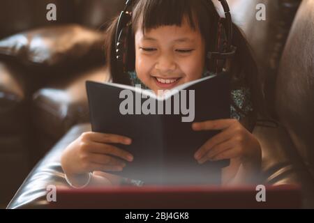 Happy little child girl lying on sofa with learning by  laptop and writing a book at home,Social distance during quarantine, Online education concept Stock Photo