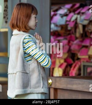 A Japanese woman praying at  the Enoshima Shrine. Ema (wood prayer and wishes plaques) can be seen in the background Stock Photo