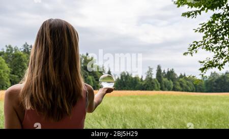 View from behind of a young woman standing in beautiful nature holding crystal globe in her hand in a conceptual image of global environmental preserv Stock Photo