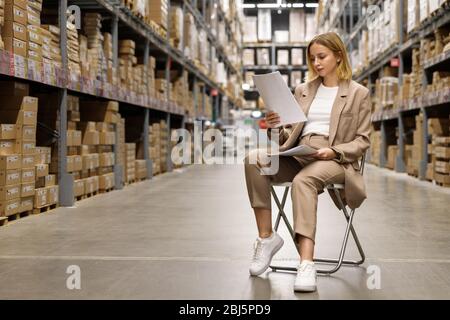Seriously business woman or supervisor in a beige suit checking documents, sitting on a chair at empty facility storage/warehouse. Data analysis, work Stock Photo