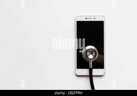 Closeup of black stethoscope on broken mobile smartphone after drop, view from above, on white background. Copy space. Smart phone with cracked screen Stock Photo