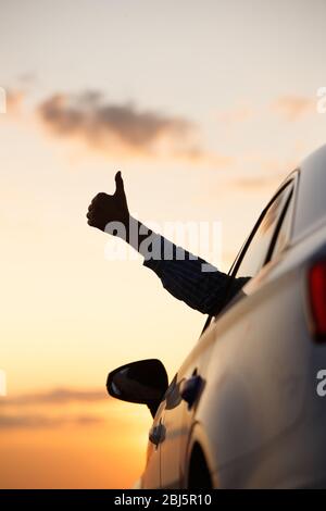 Man showing thumbs up/making Like / Ok sign with hand from car window with sunset sky, relaxing, enjoying road trip and feeling the air and freedom. T Stock Photo