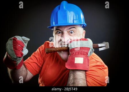 Angry worker with helmet and hummer. Mad worker biting hammer showing fist. Stock Photo