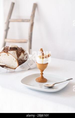 Two crackers with sesame seeds in a broken boiled egg on a wooden stand on a table with a white tablecloth and bread in the background Stock Photo