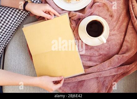 Female hands with open book, plaid, fruits and coffee on sofa. Top view Stock Photo