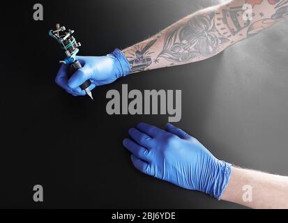 Tattooist hands in blue gloves with tattoo machine on black background Stock Photo