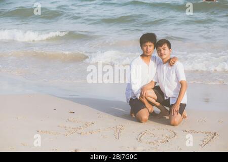 Young asian couple gay smiling romantic drawing word text love together on sand in vacation, homosexual happy and fun with love sitting on sand at the Stock Photo
