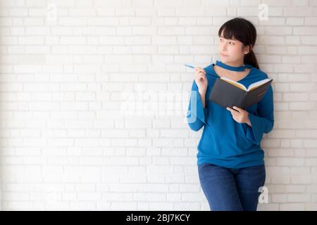 Beautiful asian woman smiling standing thinking and writing notebook on concrete cement white background at home, girl homework on book, education and Stock Photo