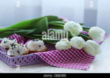 White spring tulips and delicious cupcakes in a purple tray covered with checkered napkin on a white table, close up Stock Photo
