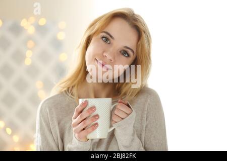 Blonde beautiful smiling girl wearing white shirt with opened eyes enjoying her cup of hot delicious coffee or tea on the light background Stock Photo