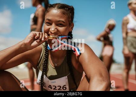 Excited female athlete biting her medal while sitting on race track with other athletes in background. Sports woman enjoying winning a medal in sprint Stock Photo