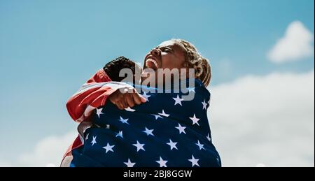 Excited american female athletes wrapped in national flag hugging each other after a winning the race. Team of USA female runners rejoicing a victory Stock Photo