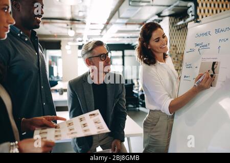 Woman giving presentation over whiteboard to team. Female explaining new marketing strategy to coworkers in office. Stock Photo