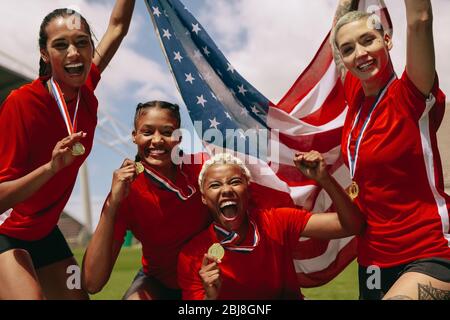 Group of female soccer team with medals holding US national flag in the field. Woman soccer team celebrating championship victory. Stock Photo