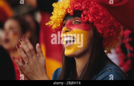 Woman sitting in stadium wearing a wig and her face painted in german flag colors applauding their team. Female from Germany in fan zone watching a so Stock Photo