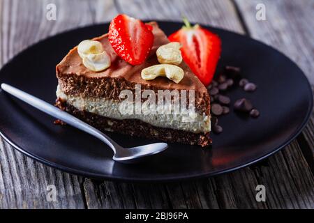 Chocolate tart of two chocolate layers: white and dark with unbaked crust of raw dates, cocoa, and nuts on a black plate with fresh strawberries and c Stock Photo