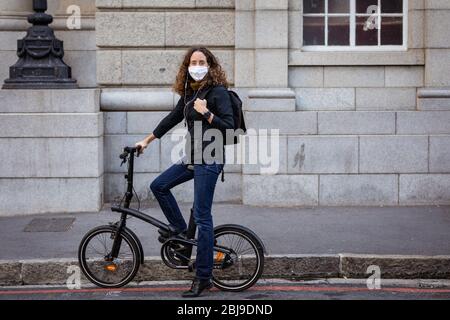 Caucasian woman wearing a protective mask and earphones, and biking in the streets Stock Photo