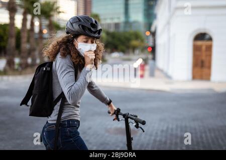 Caucasian woman wearing a protective mask and a cycling helmet in the streets, coughing in the stree Stock Photo