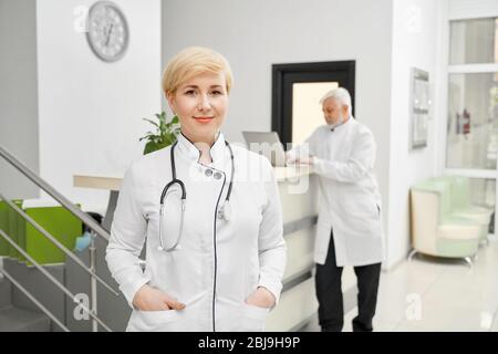 Professional therapist with stethoscope on neck posing looking at camera, eldery man using laptop on reception on background. Stock Photo
