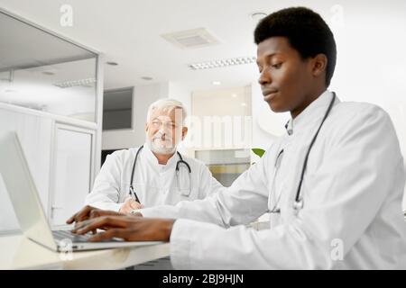 Multiracial therapists with stethoscopes on necks posing on reception in hospital. African male doctor using laptop, selective focus of caucasian elde Stock Photo