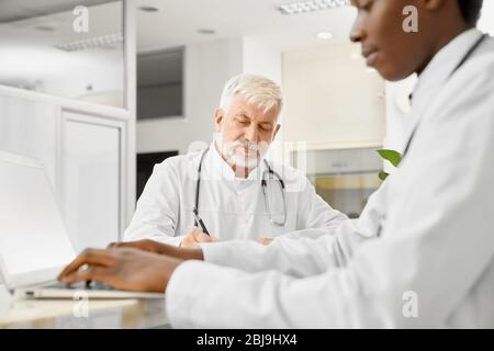 Therapists with stethoscopes on necks posing on reception in hospital. African male doctor using laptop, selective focus of caucasian eldery doctor ho Stock Photo