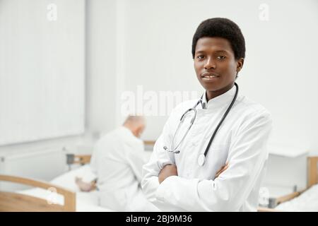 Young smiling african male doctor with stethoscope on neck looking at camera. Little female patient sitting on coach in hospital room on background, t Stock Photo