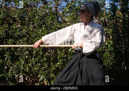 woman training Aikido in her garden during the quarantine. Weapons Aikido training - Aikido Jo kata training - Wearing black belt Aikido clothes - Tra Stock Photo