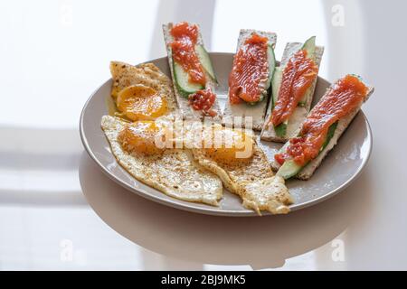 Three toasts with red fish, cucumber and three eggs on white plate over white background. Stock Photo